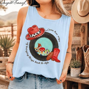 May include: A light blue sleeveless tank top featuring a graphic print of a red cowboy hat, a black tyre, and a red cowboy boot. Inside the tyre are cartoon characters. The text reads, Love has ways of bringing things back to life.
