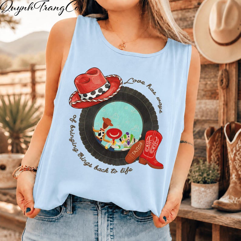 May include: A light blue sleeveless tank top featuring a graphic print of a red cowboy hat, a black tyre, and a red cowboy boot. Inside the tyre are cartoon characters. The text reads, Love has ways of bringing things back to life.