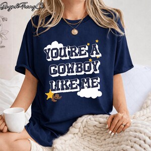 May include: A navy blue short-sleeve t-shirt featuring the white text "YOU'RE A COWBOY LIKE ME" in a bold, blocky font. The design includes white cloud shapes, small yellow stars, a yellow sheriff star badge, and a brown cowboy hat graphic.