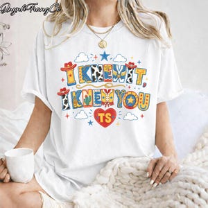 Puede incluir: Una camiseta blanca de manga corta con un estampado gr&aacute;fico colorido que incluye el texto "I KNEW IT, I KNEW YOU" y "TS" dentro de un coraz&oacute;n rojo. El dise&ntilde;o presenta sombreros de vaquero, botas, cactus, estrellas y nubes.