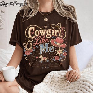 May include: A dark brown short-sleeve t-shirt featuring a graphic print with the text Cowgirl Like Me and Takes One to Know One. The design includes a red cowboy hat, a horseshoe, a rocking horse, musical notes, stars, and pink roses.