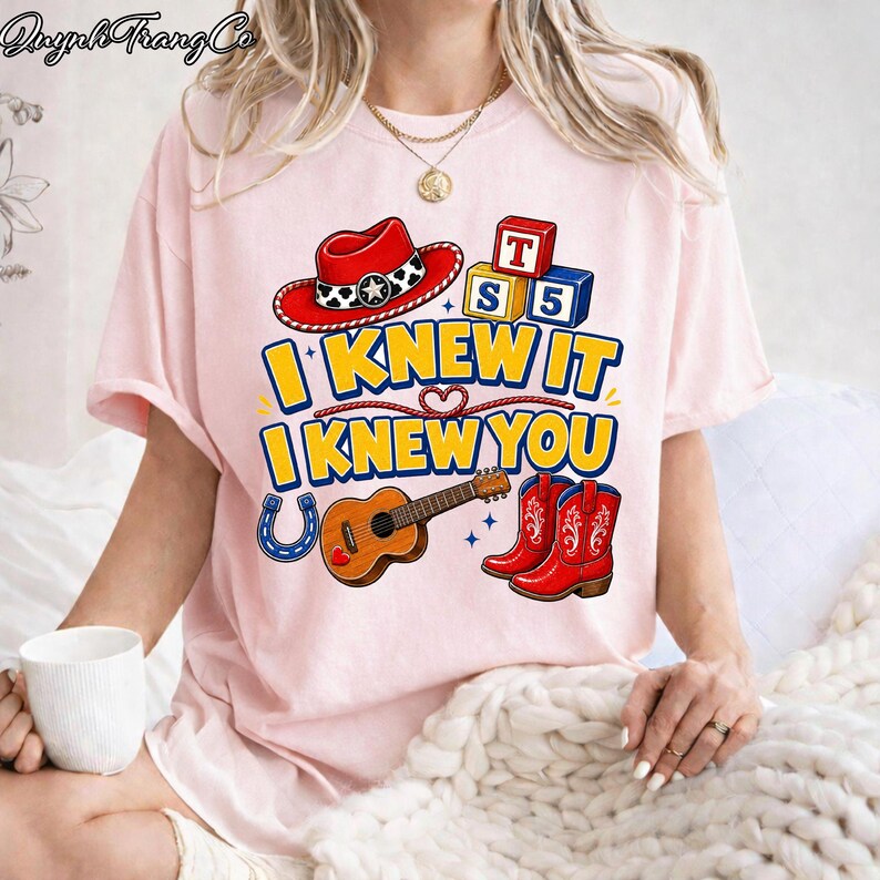 May include: A light pink t-shirt featuring a graphic print with a red cowboy hat, a brown acoustic guitar, red cowboy boots, a blue horseshoe, and alphabet blocks labeled T, S, and 5. The text reads I KNEW IT I KNEW YOU in bold yellow letters.