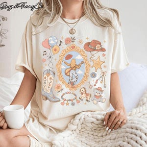 Op de afbeelding: Een cr&egrave;mekleurig T-shirt met korte mouwen en een centrale, versierde gouden spiegellijst met een figuur die gitaar speelt. De afbeelding bevat ook een cowboylaars, een hoed, een sheriffster, een teddybeer, een hobbelpaard en ballonnen.