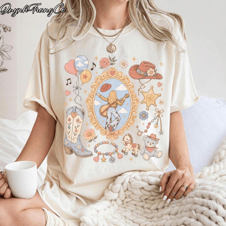 Op de afbeelding: Een cr&egrave;mekleurig T-shirt met korte mouwen en een centrale, versierde gouden spiegellijst met een figuur die gitaar speelt. De afbeelding bevat ook een cowboylaars, een hoed, een sheriffster, een teddybeer, een hobbelpaard en ballonnen.