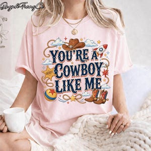 May include: A light pink t-shirt featuring a graphic print with the text "YOU'RE A COWBOY LIKE ME". The design includes a brown cowboy hat, a sheriff star badge, a rocket, a colorful ball, and cowboy boots, surrounded by rope and cloud motifs.