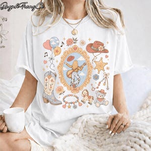 Op de afbeelding: Een wit t-shirt met korte mouwen en een kleurrijke grafische print van een centrale, sierlijke spiegellijst met een figuur met een gitaar, omringd door een cowboyhoed, een sheriffster, een westerlaars, ballonnen en een teddybeer.