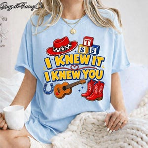 May include: A light blue t-shirt featuring a graphic print with a red cowboy hat, alphabet blocks labeled T, S, and 5, a brown acoustic guitar, and red cowboy boots. The text reads I KNEW IT I KNEW YOU in yellow letters with a blue outline.