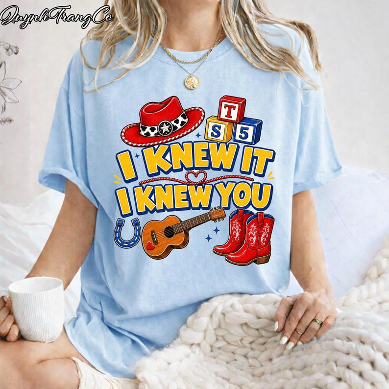 May include: A light blue t-shirt featuring a graphic print with a red cowboy hat, alphabet blocks labeled T, S, and 5, a brown acoustic guitar, and red cowboy boots. The text reads I KNEW IT I KNEW YOU in yellow letters with a blue outline.