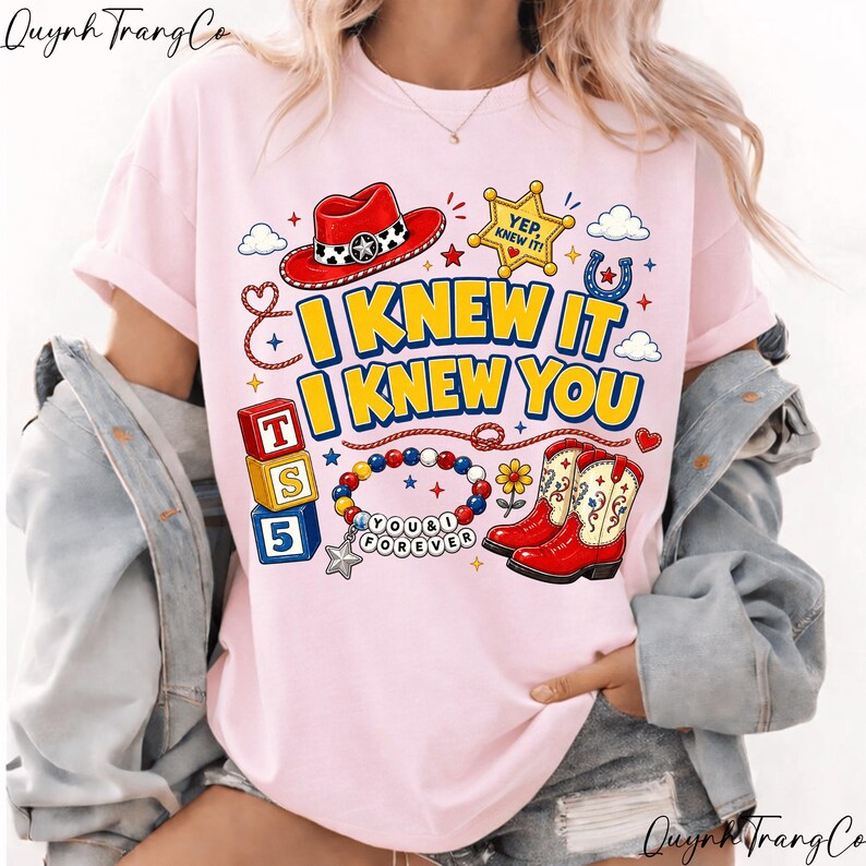 May include: A light pink t-shirt featuring a graphic print with the text I KNEW IT I KNEW YOU. The design includes a red cowboy hat, a yellow sheriff star badge, three stacked alphabet blocks, a beaded bracelet, and red cowboy boots.