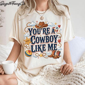 May include: A cream-colored t-shirt featuring a graphic print with the text "YOU'RE A COWBOY LIKE ME" in dark blue. The design includes a brown cowboy hat, a rocket, a sheriff star, a colorful ball, and cowboy boots, surrounded by rope accents.