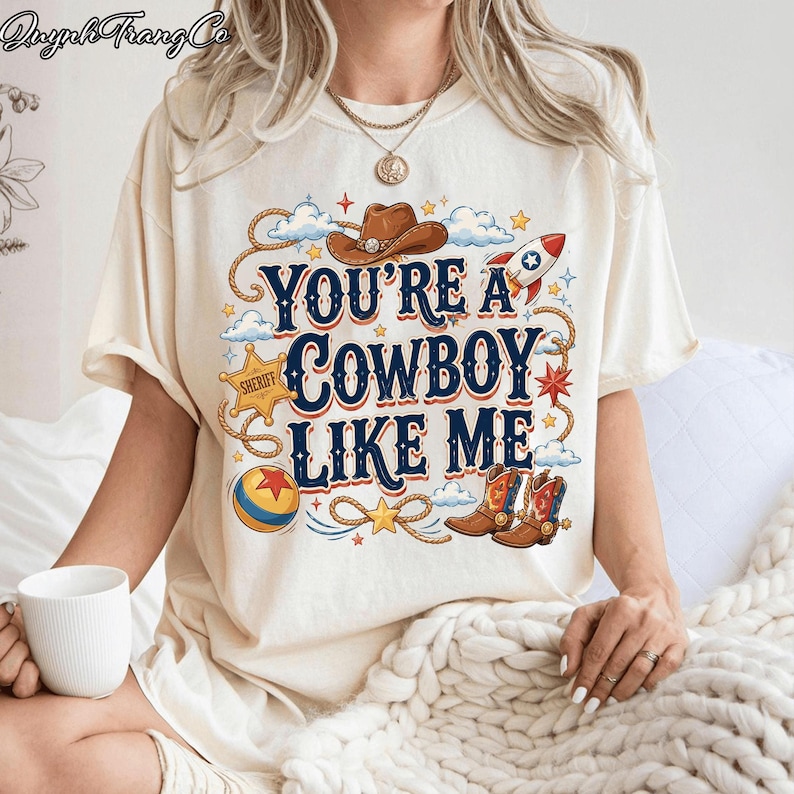 May include: A cream-colored t-shirt featuring a graphic print with the text "YOU'RE A COWBOY LIKE ME" in dark blue. The design includes a brown cowboy hat, a rocket, a sheriff star, a colorful ball, and cowboy boots, surrounded by rope accents.