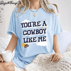 May include: A light blue short-sleeve t-shirt featuring the dark blue text "YOU'RE A COWBOY LIKE ME" in a bold font. The design includes white cloud shapes, small yellow stars, and a small brown cowboy hat icon near the bottom left corner.