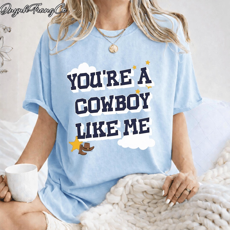 May include: A light blue short-sleeve t-shirt featuring the dark blue text "YOU'RE A COWBOY LIKE ME" in a bold font. The design includes white cloud shapes, small yellow stars, and a small brown cowboy hat icon near the bottom left corner.