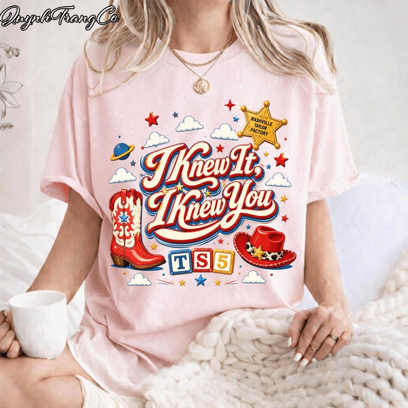 May include: A light pink t-shirt featuring a graphic print with the text "I Knew It, I Knew You" in a retro font. The design includes a red cowboy boot, a red cowboy hat, a yellow sheriff star labeled "NASHVILLE TAYLOR FACTORY", and "TS5" blocks.