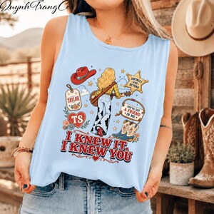 May include: A light blue sleeveless tank top featuring a graphic print of a person with blonde hair playing a guitar, wearing cow print trousers. The design includes a red cowboy hat, a star, a heart, and boots, with the text "I KNEW IT, I KNEW YOU."