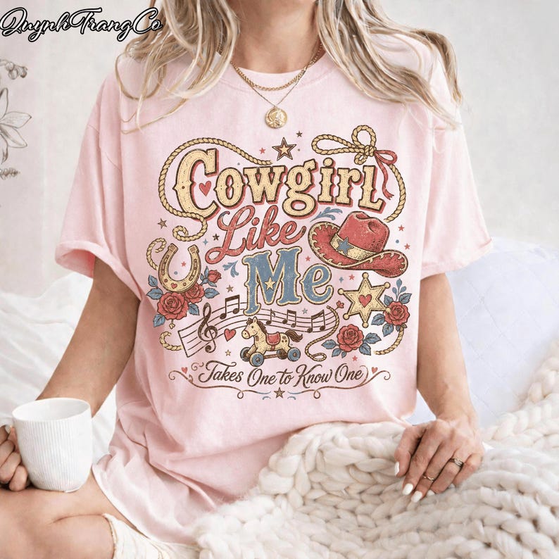 May include: A light pink t-shirt featuring a graphic print with the text Cowgirl Like Me Takes One to Know One. The design includes a lasso, a cowboy hat, a star, a rocking horse, musical notes, and red roses in a vintage-inspired style.