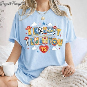 Puede incluir: Una camiseta azul claro con un estampado gr&aacute;fico colorido con el texto "I KNEW IT, I KNEW YOU" y "TS" dentro de un coraz&oacute;n rojo. El dise&ntilde;o incluye ilustraciones de tem&aacute;tica western como sombreros de vaquero, botas, cactus y estrellas.