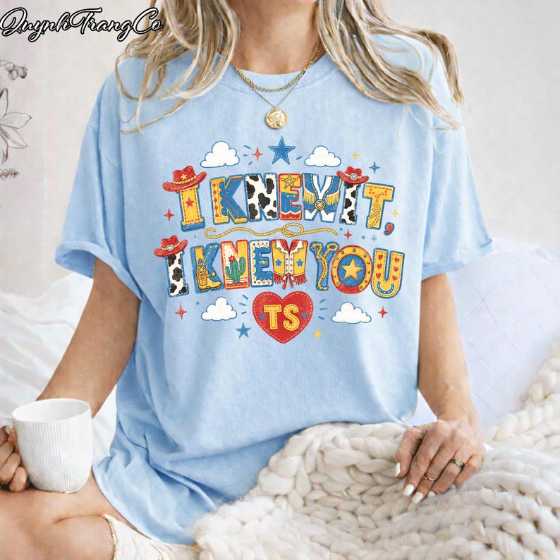 Puede incluir: Una camiseta azul claro con un estampado gr&aacute;fico colorido con el texto "I KNEW IT, I KNEW YOU" y "TS" dentro de un coraz&oacute;n rojo. El dise&ntilde;o incluye ilustraciones de tem&aacute;tica western como sombreros de vaquero, botas, cactus y estrellas.