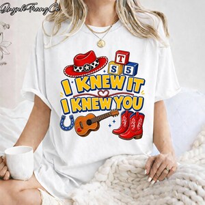 May include: A white short-sleeve t-shirt featuring a graphic print with a red cowboy hat, alphabet blocks, a brown acoustic guitar, a blue horseshoe, and red cowboy boots. The text reads I KNEW IT I KNEW YOU in bold yellow and blue letters.