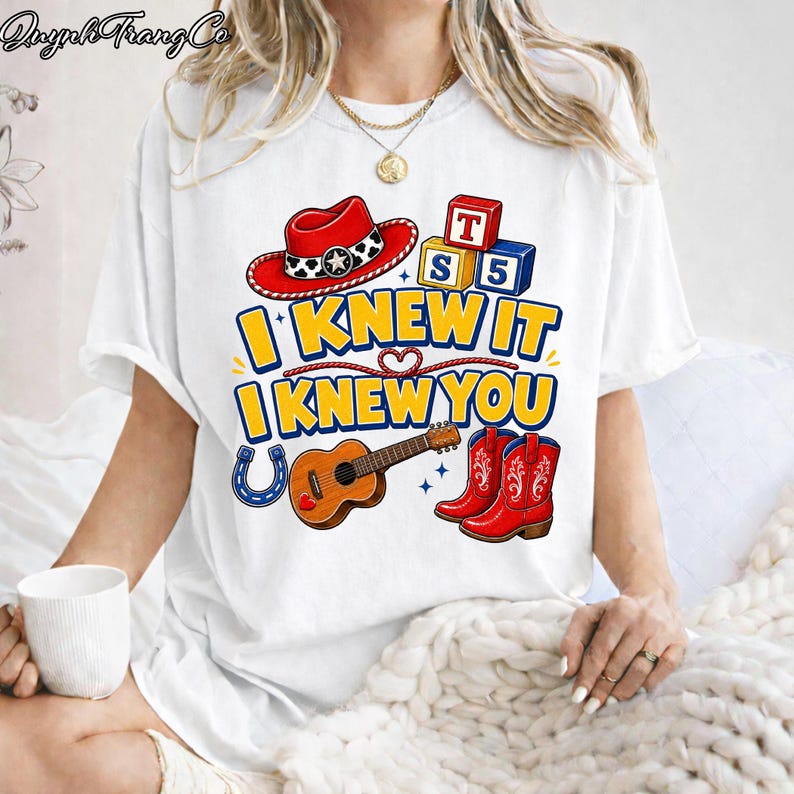 May include: A white short-sleeve t-shirt featuring a graphic print with a red cowboy hat, alphabet blocks, a brown acoustic guitar, a blue horseshoe, and red cowboy boots. The text reads I KNEW IT I KNEW YOU in bold yellow and blue letters.