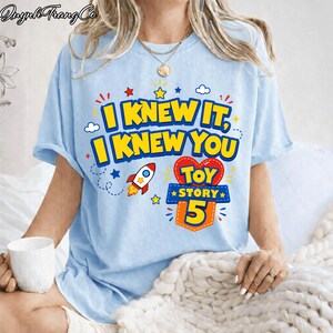 Pu&ograve; includere: Una t-shirt azzurro chiaro con una stampa grafica che riporta il testo I KNEW IT, I KNEW YOU e TOY STORY 5. Il design include un piccolo razzo, una forma a cuore rossa e stelle gialle. La t-shirt ha maniche corte e un girocollo.