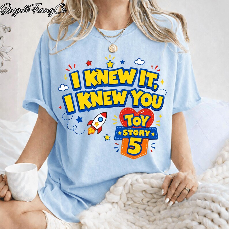 Pu&ograve; includere: Una t-shirt azzurro chiaro con una stampa grafica che riporta il testo I KNEW IT, I KNEW YOU e TOY STORY 5. Il design include un piccolo razzo, una forma a cuore rossa e stelle gialle. La t-shirt ha maniche corte e un girocollo.