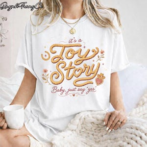 Puede incluir: Una camiseta blanca de manga corta con un dise&ntilde;o gr&aacute;fico que incluye el texto "it's a Toy Story" escrito en una fuente que simula una cuerda, acompa&ntilde;ado de la frase "Baby, just say 'yes'" e ilustraciones de una herradura, flores y un sombrero de vaquero.