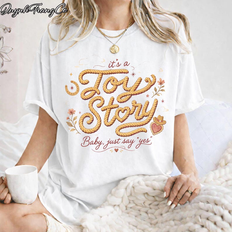 Puede incluir: Una camiseta blanca de manga corta con un dise&ntilde;o gr&aacute;fico que incluye el texto "it's a Toy Story" escrito en una fuente que simula una cuerda, acompa&ntilde;ado de la frase "Baby, just say 'yes'" e ilustraciones de una herradura, flores y un sombrero de vaquero.