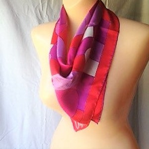 May include: A vibrant silk scarf with a geometric pattern in shades of red, pink, and purple. The scarf features a bold, square design with a contrasting red border. The scarf is draped over a mannequin.