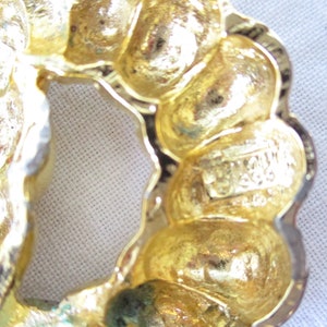 May include: Close-up of a gold-toned vintage brooch with a floral design. The brooch has a textured surface and a scalloped edge. The maker's mark is visible on the back. A classic piece of jewelry.