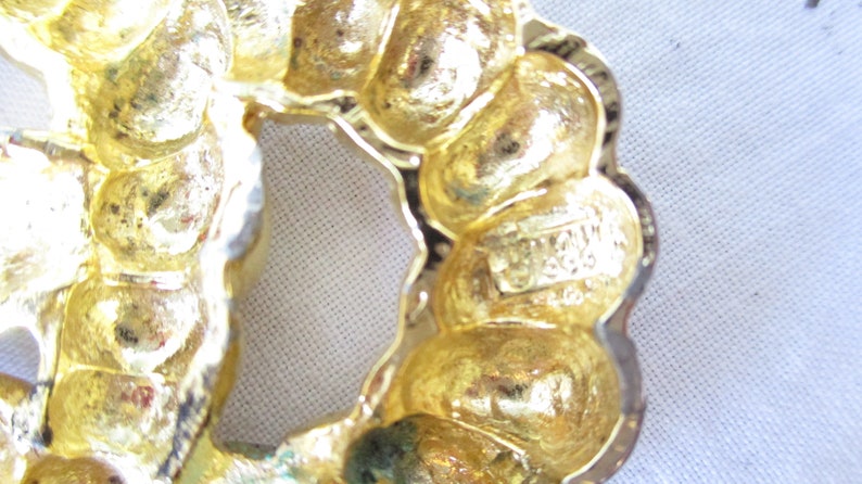 May include: Close-up of a gold-toned vintage brooch with a floral design. The brooch has a textured surface and a scalloped edge. The maker's mark is visible on the back. A classic piece of jewelry.