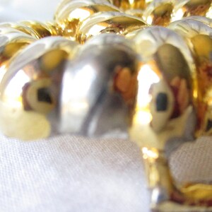 May include: Close-up of a gold-colored, decorative object with a repeating, ribbed design. The object has a shiny, reflective surface, with hints of silver and gold. The background is a soft, white fabric.
