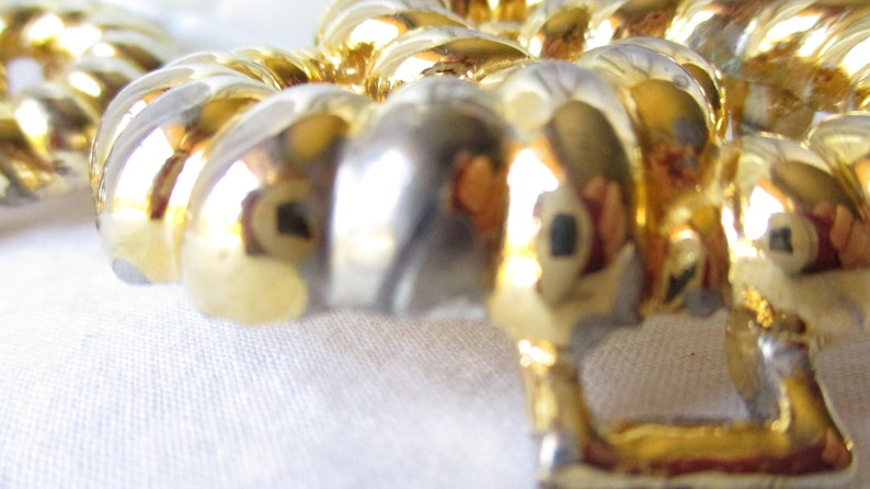 May include: Close-up of a gold-colored, decorative object with a repeating, ribbed design. The object has a shiny, reflective surface, with hints of silver and gold. The background is a soft, white fabric.