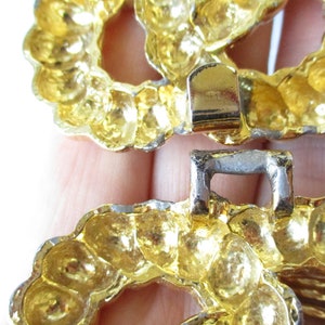 May include: Close-up of a gold-toned metal belt buckle. The buckle is composed of two interlocking circular pieces with a textured, beaded design. The clasp is silver-colored.