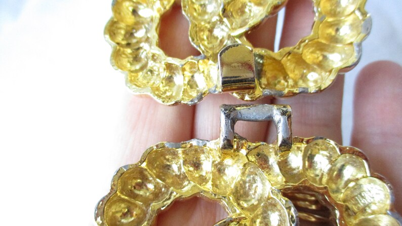 May include: Close-up of a gold-toned metal belt buckle. The buckle is composed of two interlocking circular pieces with a textured, beaded design. The clasp is silver-colored.