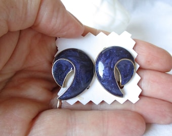 May include: A pair of vintage clip-on earrings. Each earring features a swirling, navy blue enamel design with gold-tone trim. The earrings are displayed on a white, scalloped card, held by a hand.