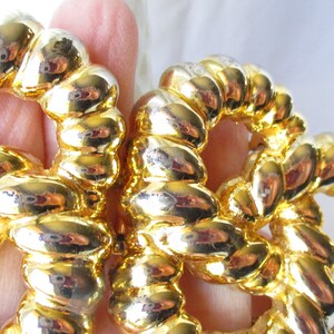 May include: Close-up of a pair of gold-colored, rope-style earrings. The earrings are made of a shiny, metallic material and have a circular design. The rope detailing is visible, and the earrings reflect light, creating a bright appearance.