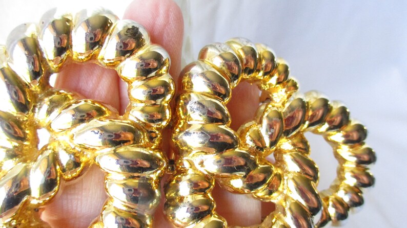 May include: Close-up of a pair of gold-colored, rope-style earrings. The earrings are made of a shiny, metallic material and have a circular design. The rope detailing is visible, and the earrings reflect light, creating a bright appearance.