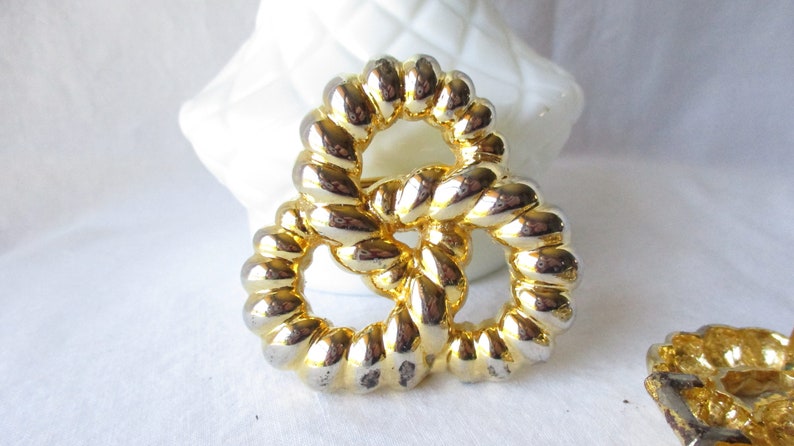 May include: A gold-toned metal brooch with a pretzel-like design. The brooch features a textured, rope-like pattern and a shiny finish. The jewelry is set against a white background, highlighting its intricate details.