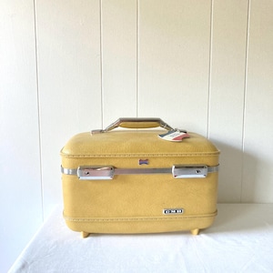 May include: A mustard yellow hard-shell train case with a textured finish, featuring a silver-tone metal handle, two silver-tone latches, and a small rectangular metal plate on the front with the letters CMB printed in white on a black background.
