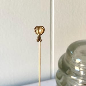 May include: A gold-colored hat pin with a heart-shaped design and a small pearl in the center. The pin is slender and has a long, straight shaft. The pin is likely vintage or antique.