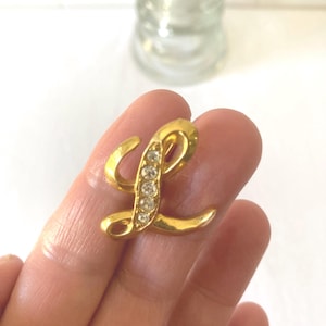 May include: A gold-colored letter "L" brooch with clear rhinestones. The brooch is in the shape of the letter "L" and has a shiny finish. The rhinestones are set in a vertical line along the letter's curve. The brooch is held in a hand.