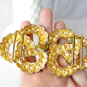 May include: A gold-toned metal belt buckle with an oval shape. The buckle is composed of two connected halves, each featuring a decorative, beaded border. The design includes a central, openwork pattern and a clasp mechanism.