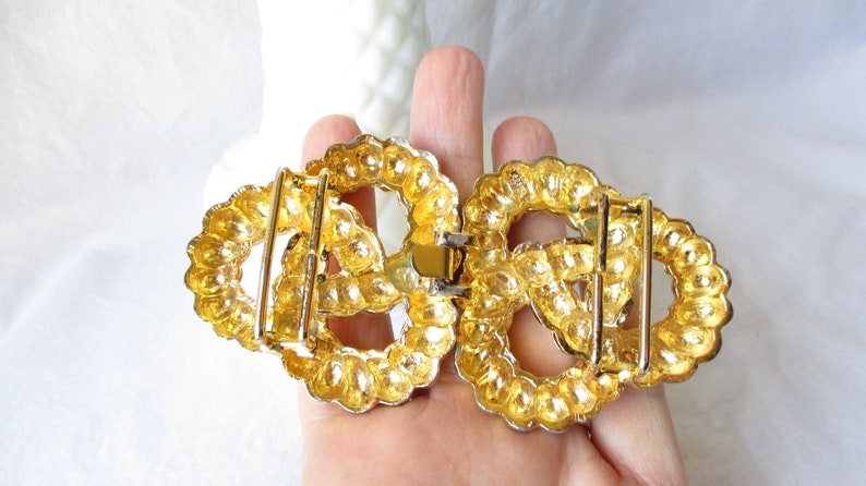 May include: A gold-toned metal belt buckle with an oval shape. The buckle is composed of two connected halves, each featuring a decorative, beaded border. The design includes a central, openwork pattern and a clasp mechanism.