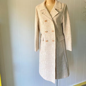May include: A double-breasted, knee-length coat in a light beige color featuring a textured, wavy vertical line pattern. The coat has a notched lapel, long sleeves, two flap pockets at the waist, and four round buttons on the front closure.