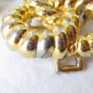 May include: Close-up of a gold-toned, circular buckle with a textured, rope-like design. The buckle has a square clasp and is set against a white background. The metal has areas of silver and gold.