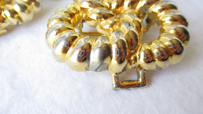 May include: Close-up of a gold-toned, circular buckle with a textured, rope-like design. The buckle has a square clasp and is set against a white background. The metal has areas of silver and gold.