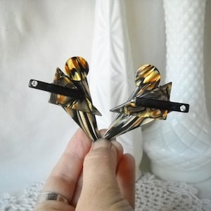 May include: A pair of vintage geometric earrings. The earrings feature a black bar with a small rhinestone, a triangle, and a circle with a brown, black, and cream marbled pattern. The earrings are held in a hand.