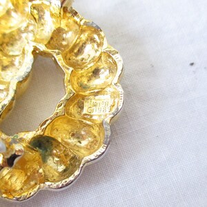 May include: Close-up of a gold-toned, textured brooch with a circular design. The brooch has a silver-colored pin and clasp. The brooch is marked with the text "&copy;1989".