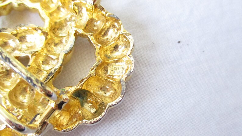 May include: Close-up of a gold-toned, textured brooch with a circular design. The brooch has a silver-colored pin and clasp. The brooch is marked with the text "&copy;1989".